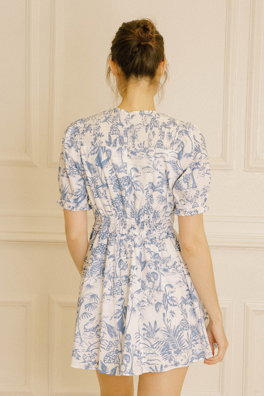 Back view of a woman wearing a blue and white tropical print mini dress against a beige wall.