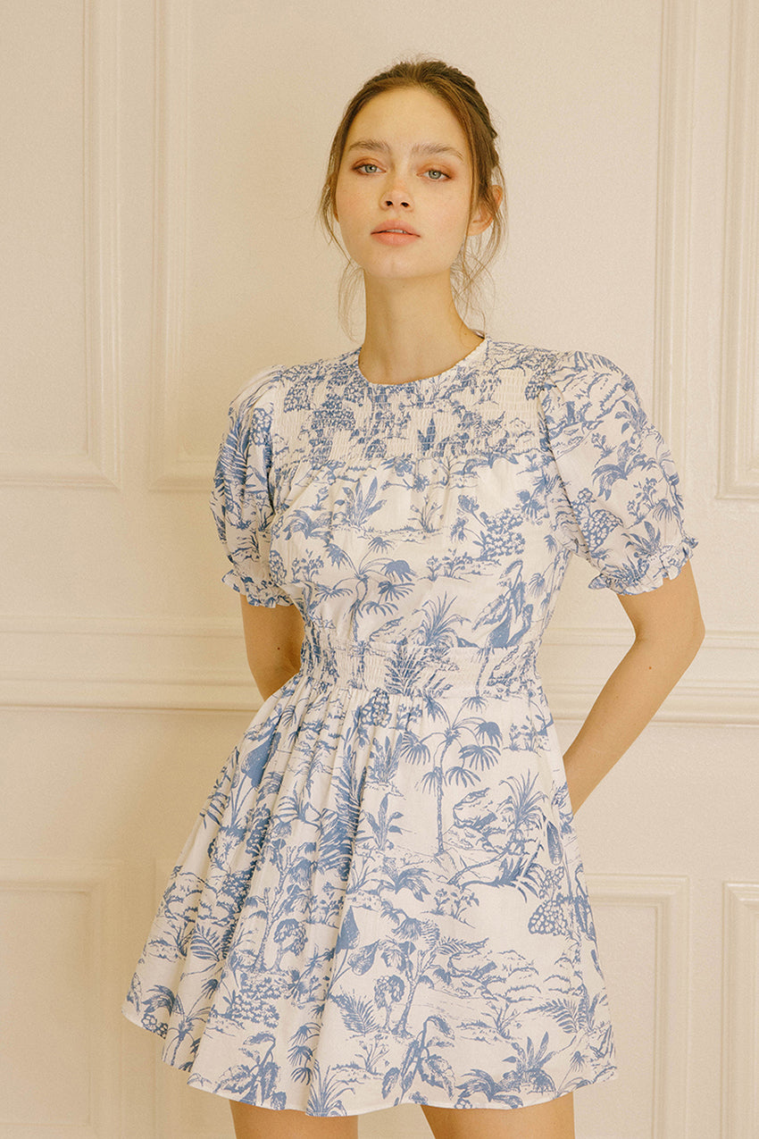 Woman wearing a blue and white tropical print mini dress against a beige wall.