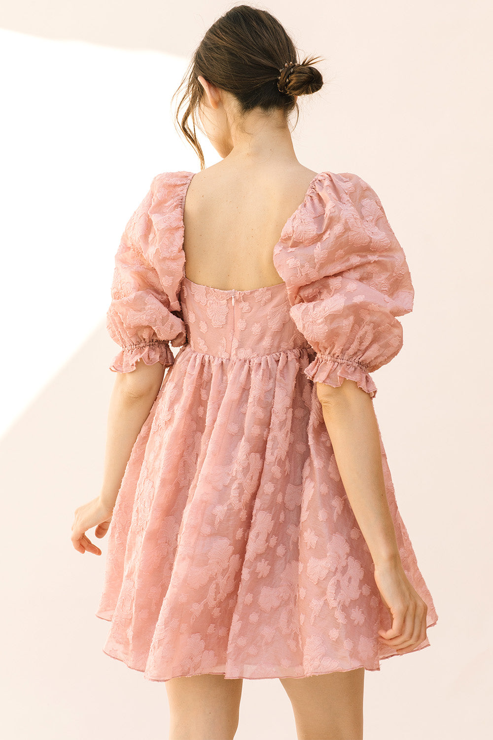 Back view of a woman wearing a pink dress with puff sleeves and an invisible zipper against a beige background