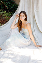 Woman in a light blue floral dress sitting on the floor against a white curtain with greenery in the background