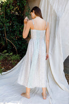 Back view of a woman in a light blue floral dress standing against a white curtain with greenery in the background