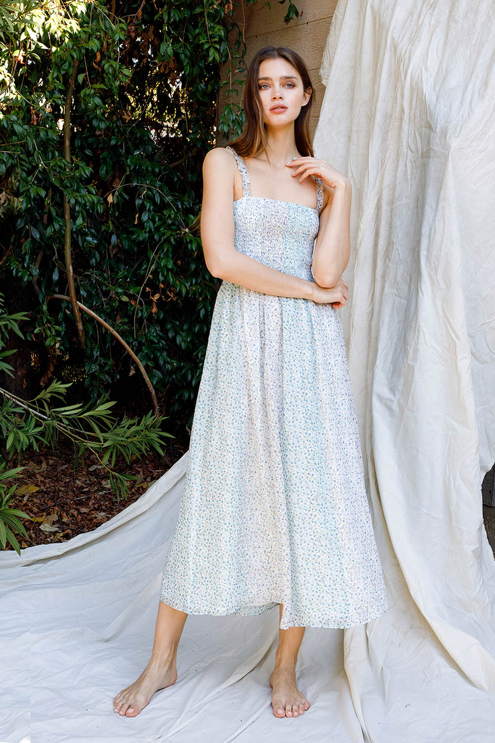 Woman in a light blue floral dress standing against a white curtain with greenery in the background