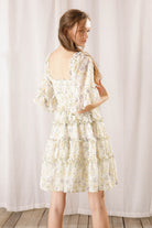 Back view of a woman wearing an ivory floral dress against a white curtain background