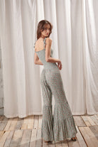 Back view of a woman wearing a green jumpsuit with a floral pattern standing in front of white curtains.