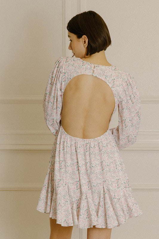 Back view of a woman wearing a pink dress with a floral pattern, open back and upper back buttons for closure against a neutral background