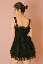 Back of a woman wearing a black tulle tiered dress against a pink background