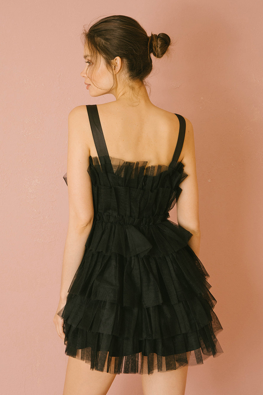 Back of a woman wearing a black tulle tiered dress against a pink background
