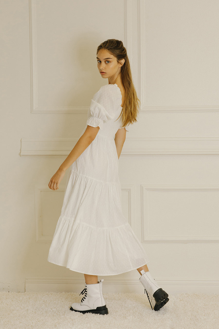 Side view of a woman wearing a white dress with puffed sleeves against a light-colored paneled wall.